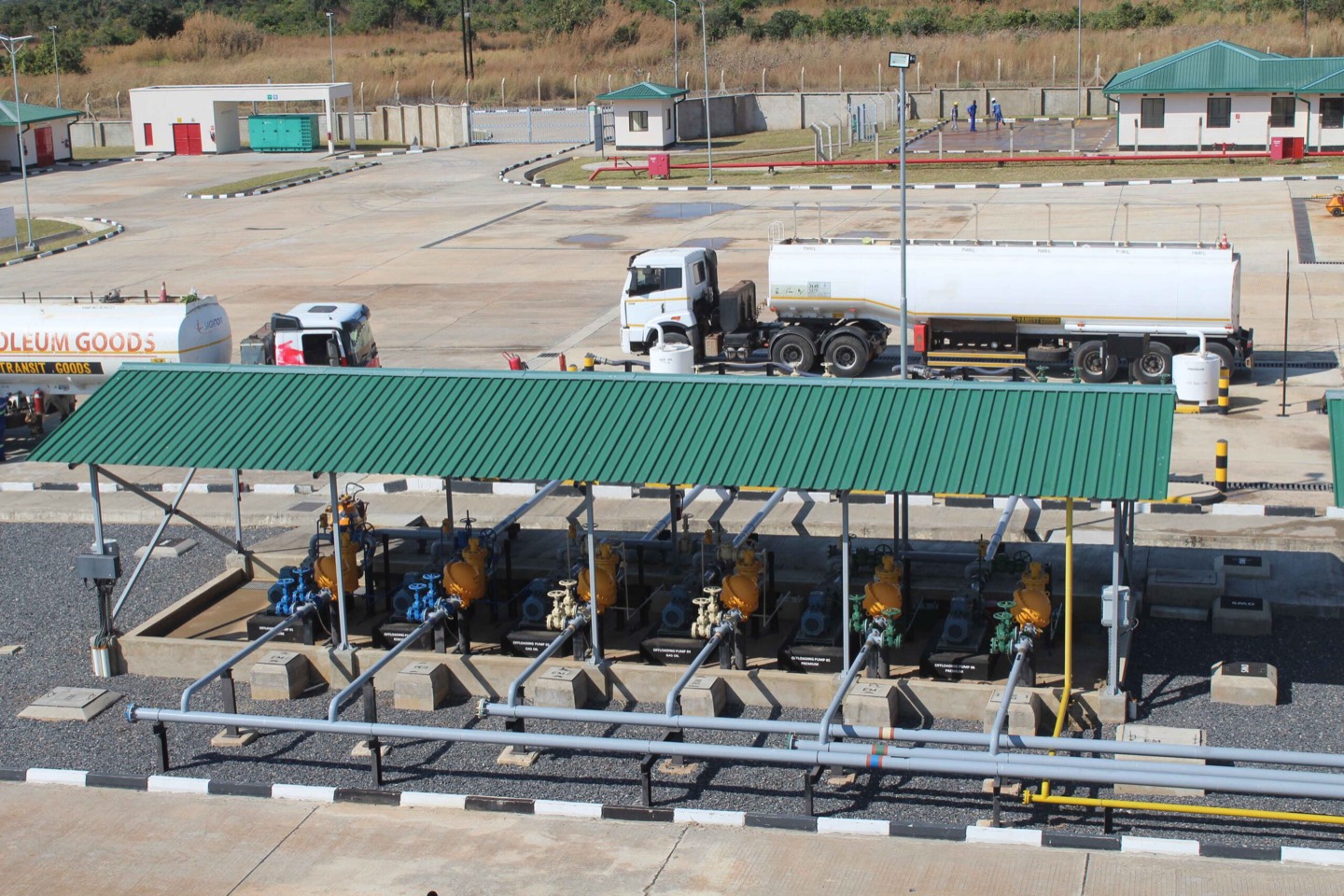Fuel depot loading gantry: loading arms and valves under a green canopy, with road tankers on the apron behind.